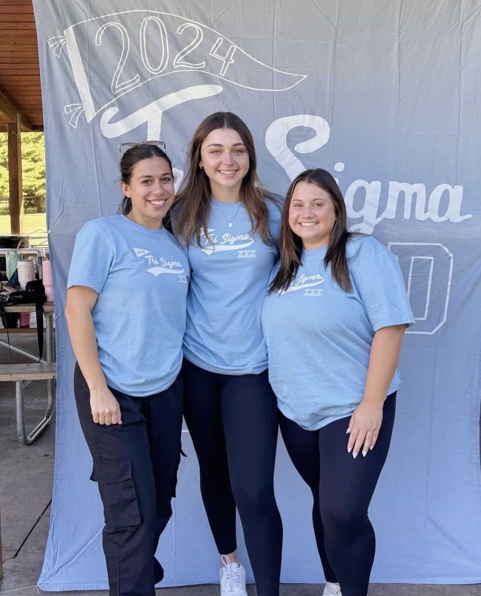 3 friends wearing the field day shirts, a  blue tee shirt with white lettering, they are standing in front of a tri sigma banner