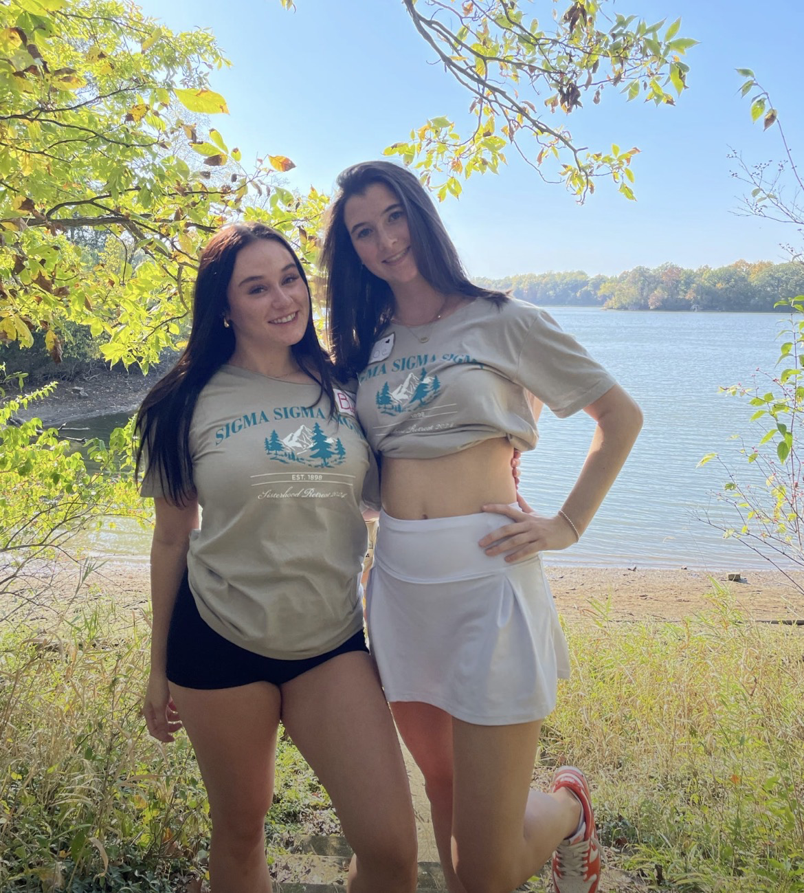  2 friends wearing the camping retreat beige tee shirts with a blue and white trees and mountains design, they stand infront of a lake for the photo 