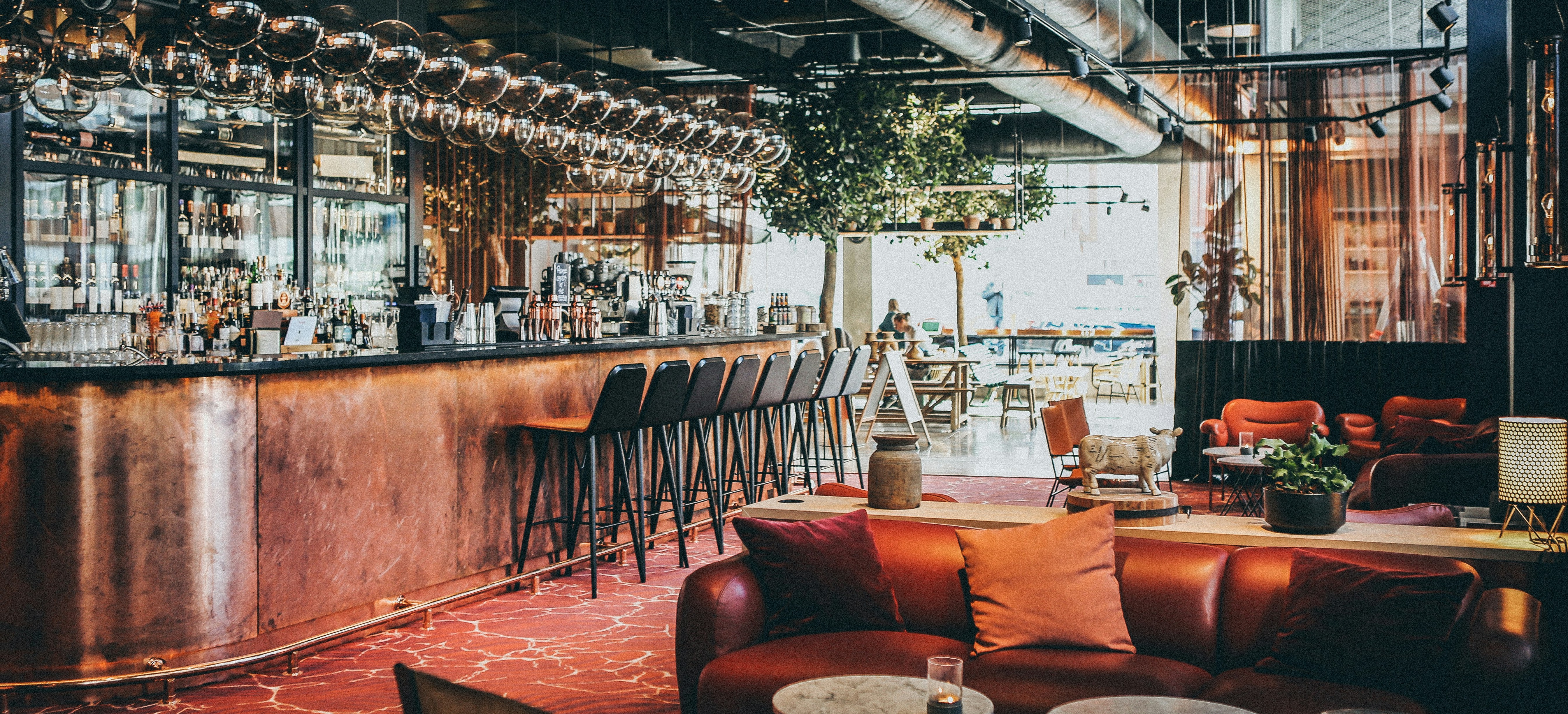  an image of the inside of sugar shot showen as fancy bar lounge area  with seating, couches and a bar in the background, dim lighting and large windows letting in the light. green vines and rustic decor and bottles decorate the lounge