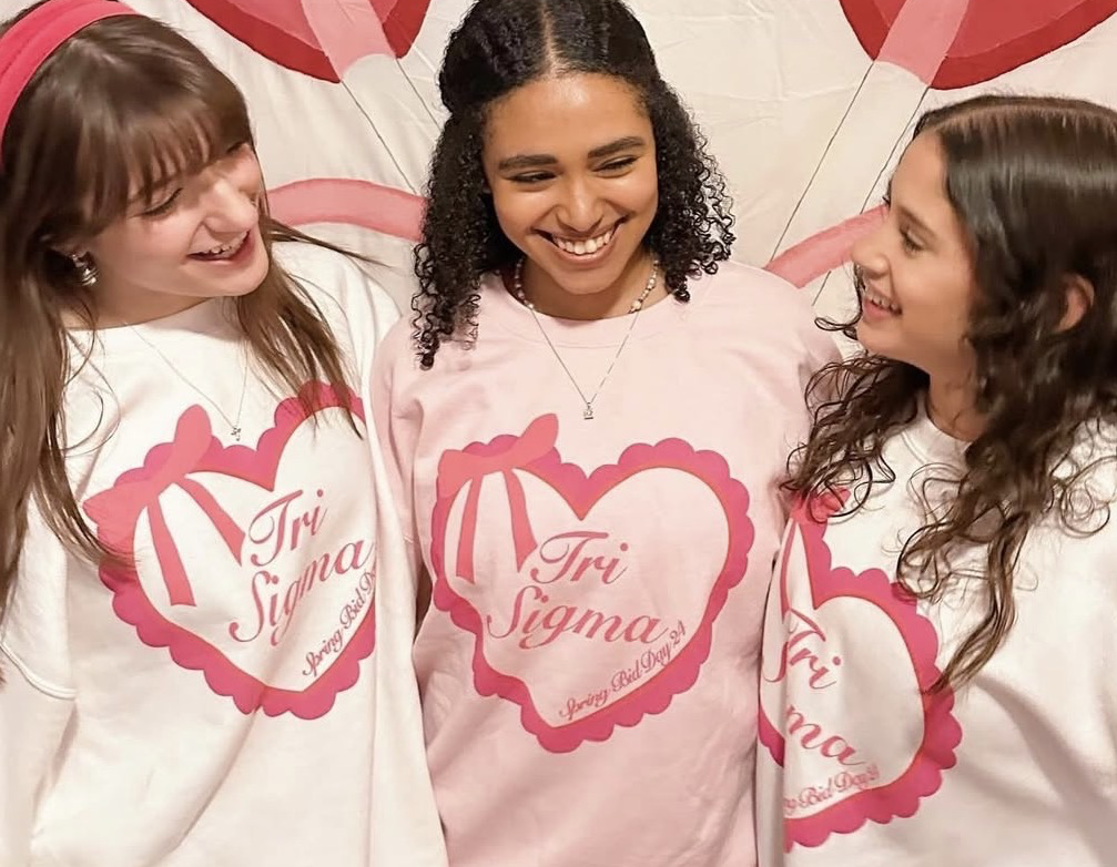 me along with 2 other friends wearing the winter bid day heart sweatshirts in pink and white with a tri sigma poster as the background of the photo.